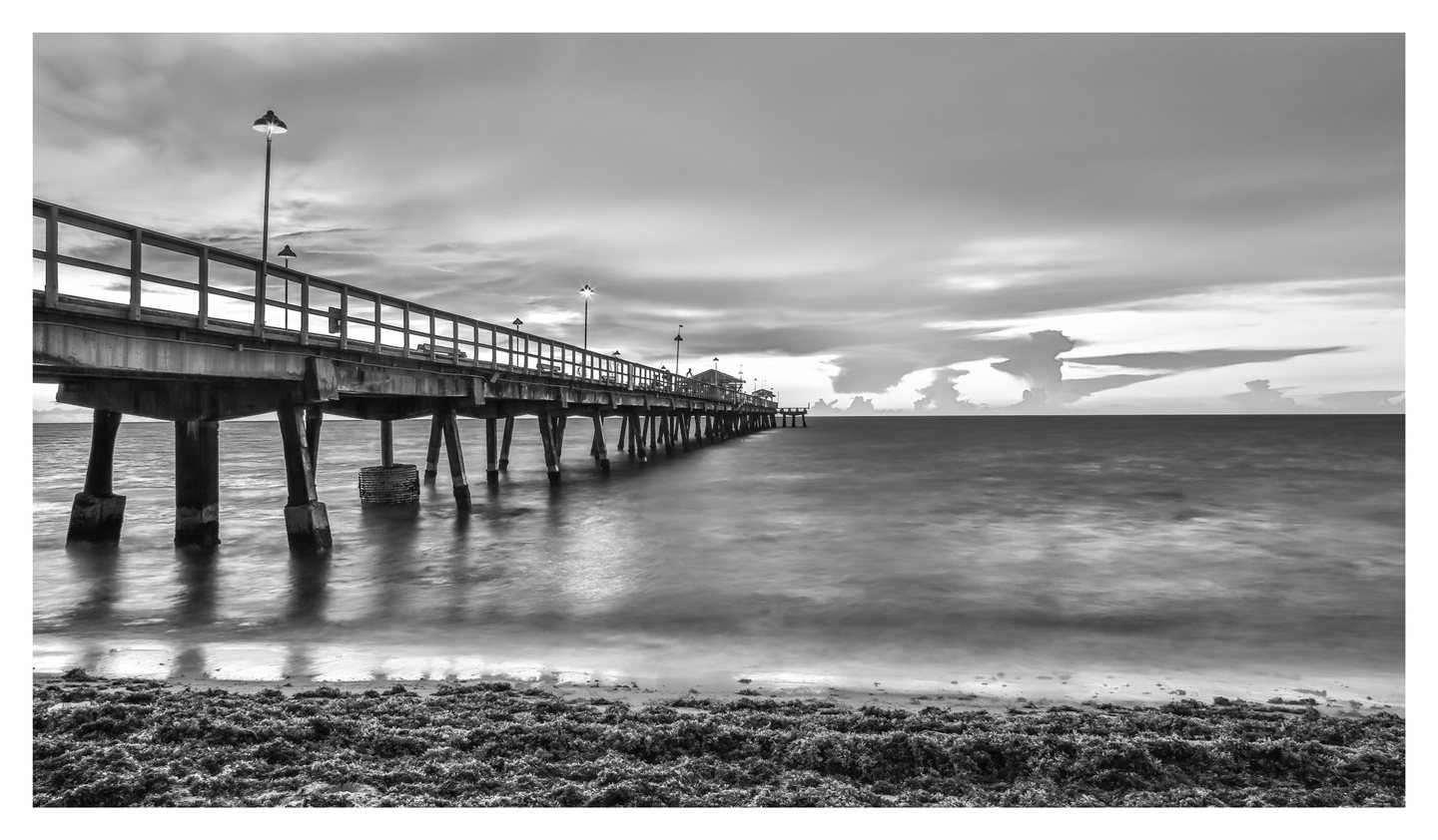 Black-and-white photograph of a lamp-lit pier extending from the left over softly blurred ocean waves beneath layered clouds, with seaweed covering the foreground beach.