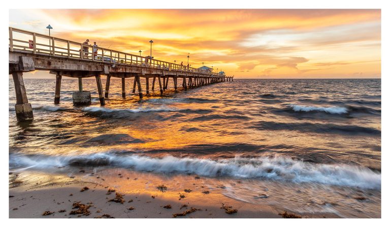 A fishing pier extends over ocean waves beneath a glowing orange and gold sky, with white surf washing onto seaweed-strewn sand.