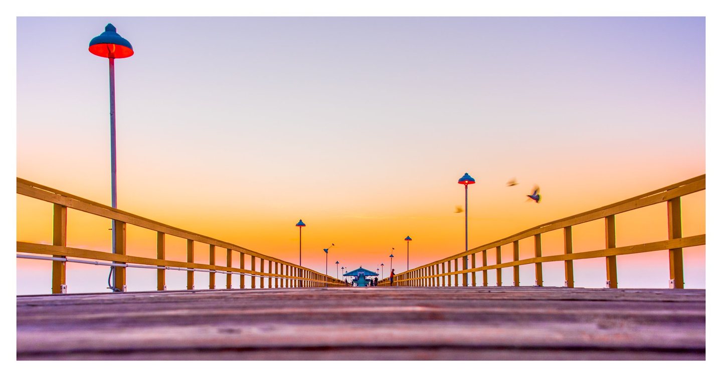 A low-angle view along a wooden pier shows yellow railings, glowing lamps, distant people, and birds flying across an orange and lavender sky.
