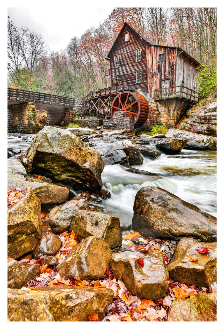 A weathered wooden mill with a rust-red waterwheel stands beside a bridge over a rocky stream, surrounded by sparse autumn woodland and fallen orange leaves.
