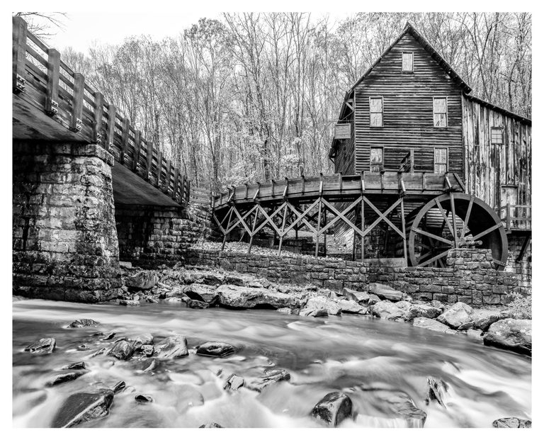 Black-and-white photograph of a wooden gristmill and large waterwheel beside a rocky stream, with a bridge on the left and woodland behind.