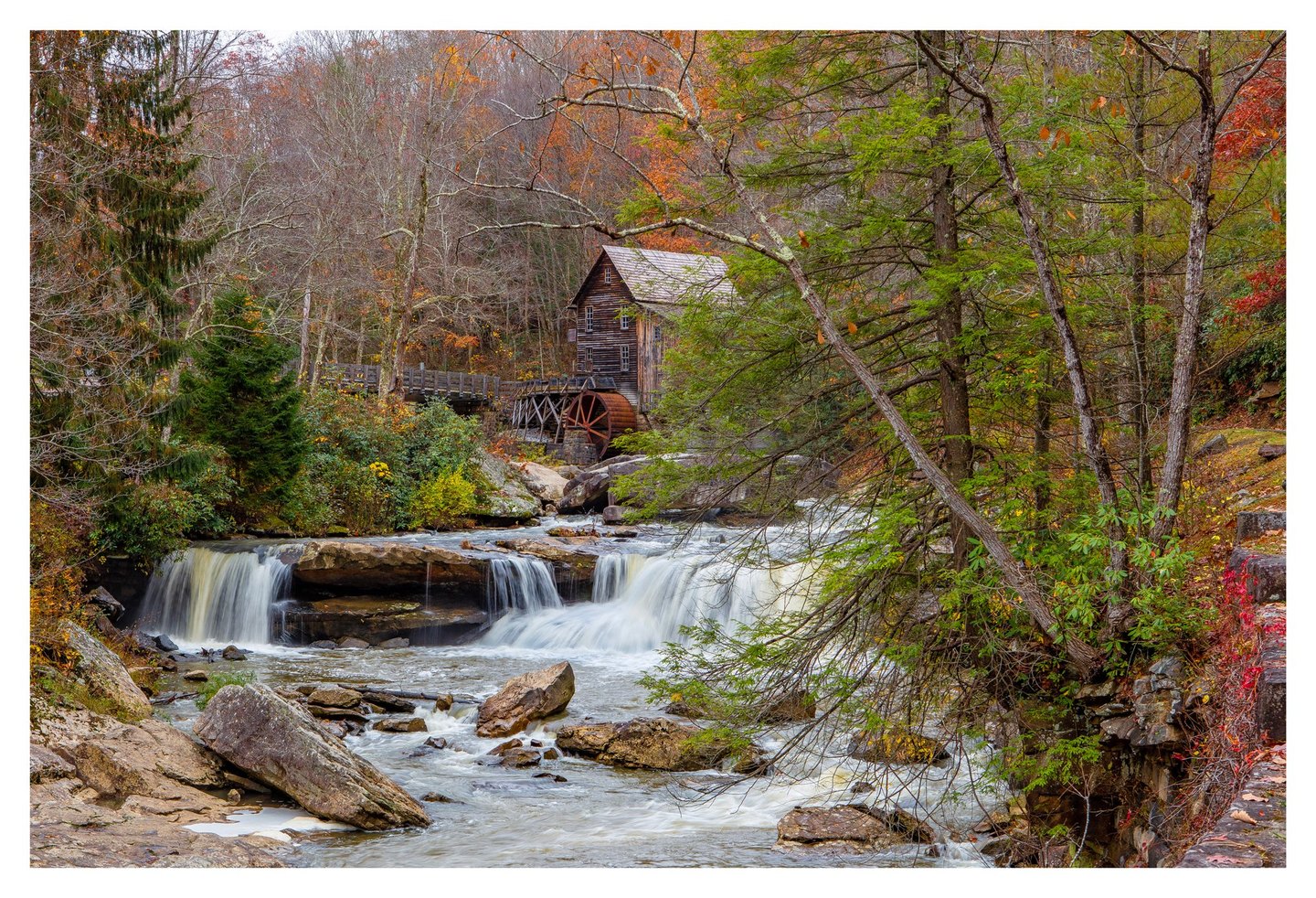 A wooden watermill with a rust-colored wheel stands among autumn trees above a rocky stream cascading over broad stone ledges.