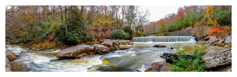 A panoramic view of a rocky river flowing over a low weir and around large boulders, bordered by autumn woodland and a stone retaining wall.