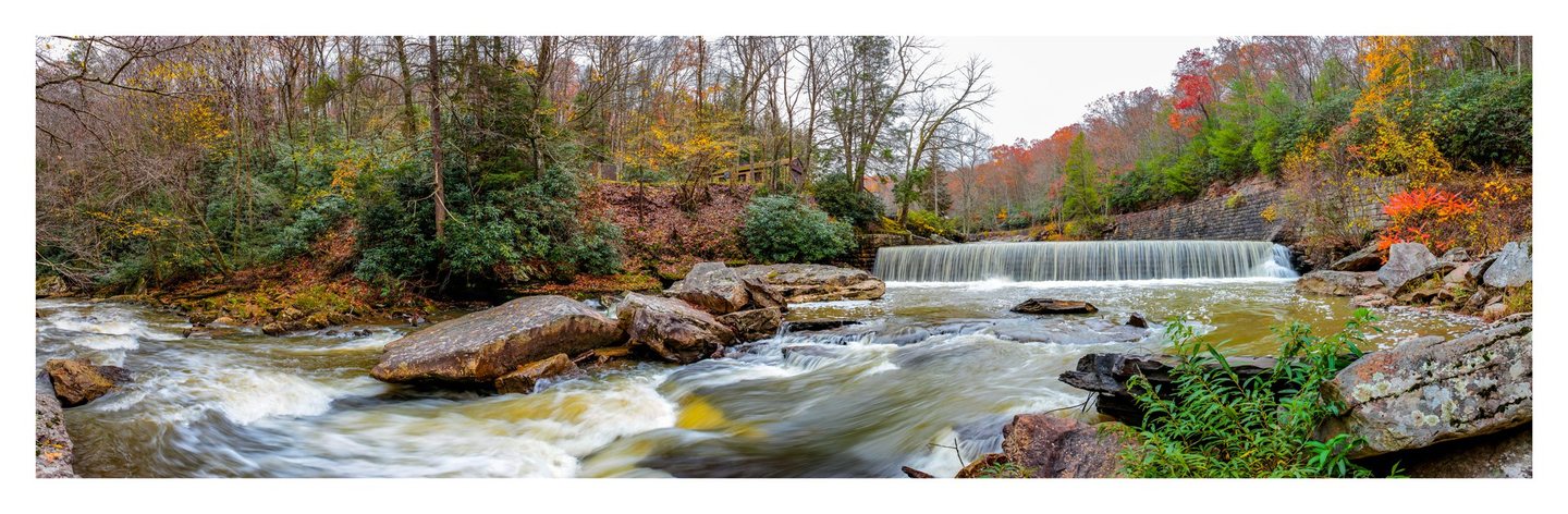 A panoramic view of a rocky river flowing over a low weir and around large boulders, bordered by autumn woodland and a stone retaining wall.