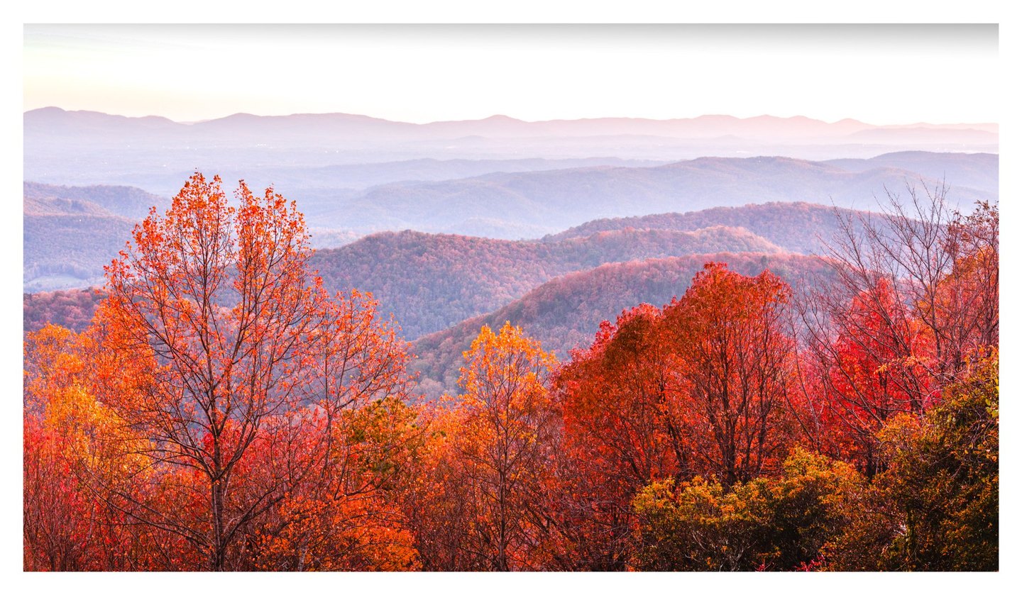 Vivid red and orange autumn trees overlook layers of forested mountain ridges fading into lavender haze beneath a pale sky.