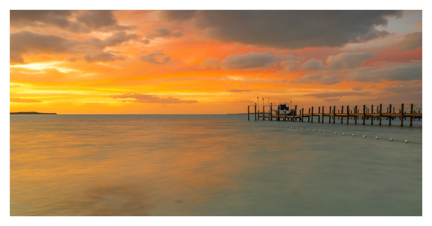 An orange and gold sunset reflects across calm coastal water beside a wooden dock with moored boats and a curving line of white buoys.