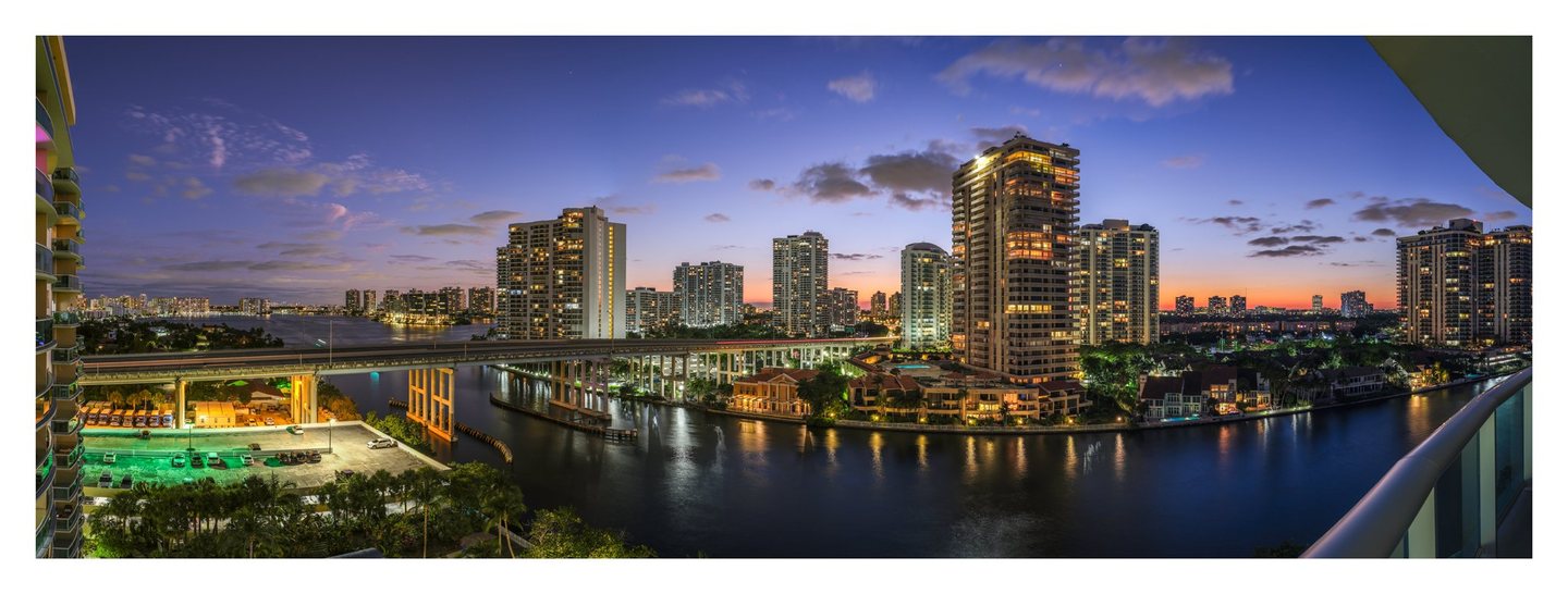 Panoramic balcony view of waterfront high-rises and a bridge at dusk, with illuminated windows reflecting on dark water beneath a blue and orange sky.