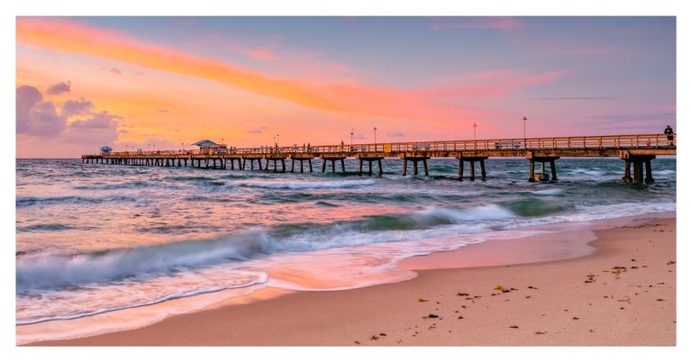 Waves wash onto a sandy beach beneath a long pier with scattered people and lamps, while a pink and orange sky reflects across the water.
