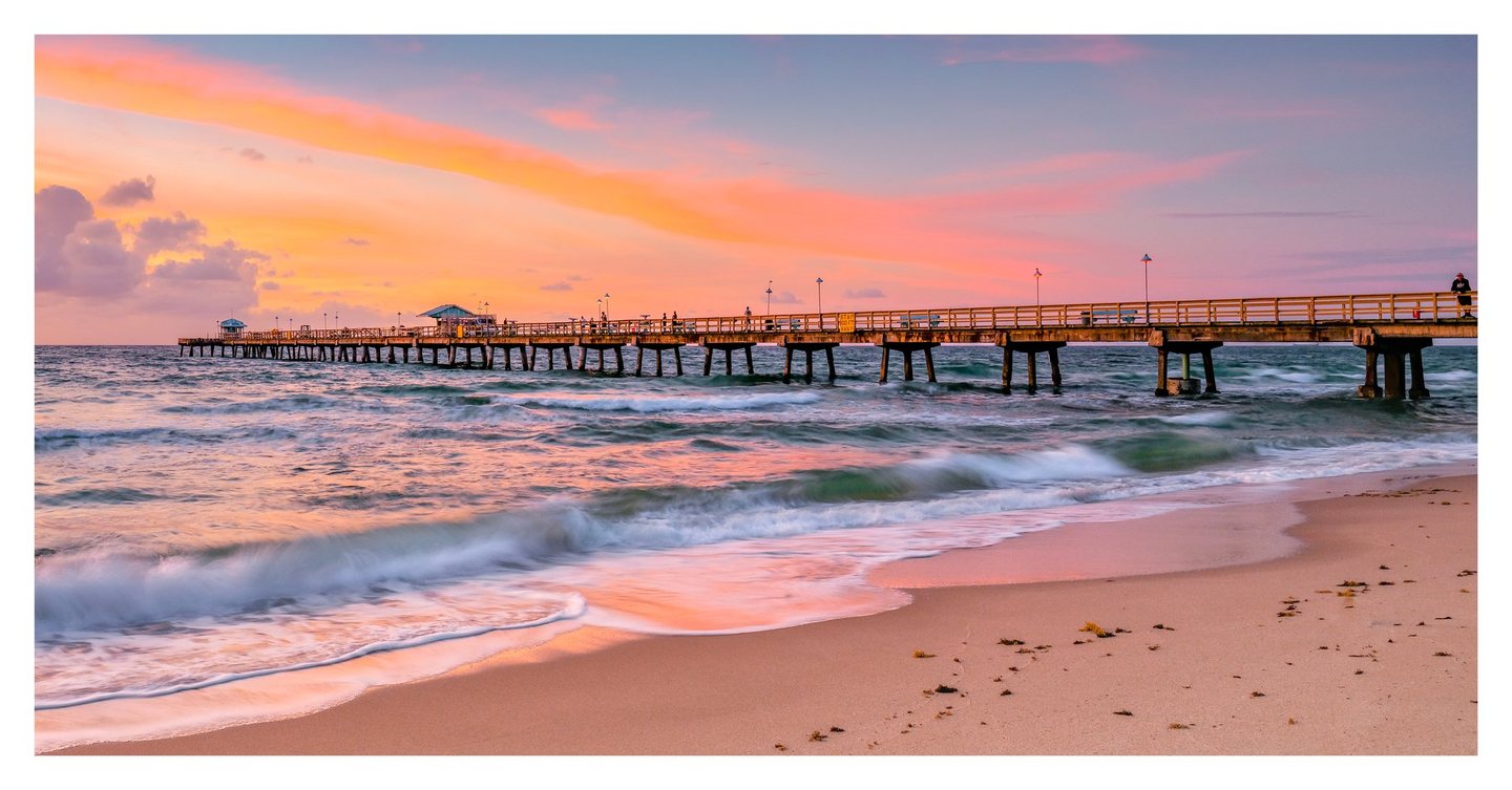 Waves wash onto a sandy beach beneath a long pier with scattered people and lamps, while a pink and orange sky reflects across the water.