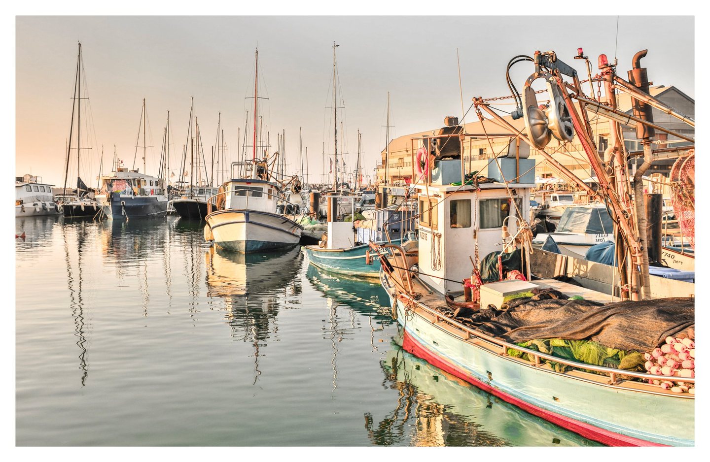 Fishing boats and sailboats sit moored in a sunlit harbor, with nets and metal rigging on a pale turquoise boat in the foreground and masts reflected in calm water.