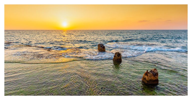 A low sun casts golden light across the sea as small waves curl around three weathered concrete remnants in clear coastal shallows.