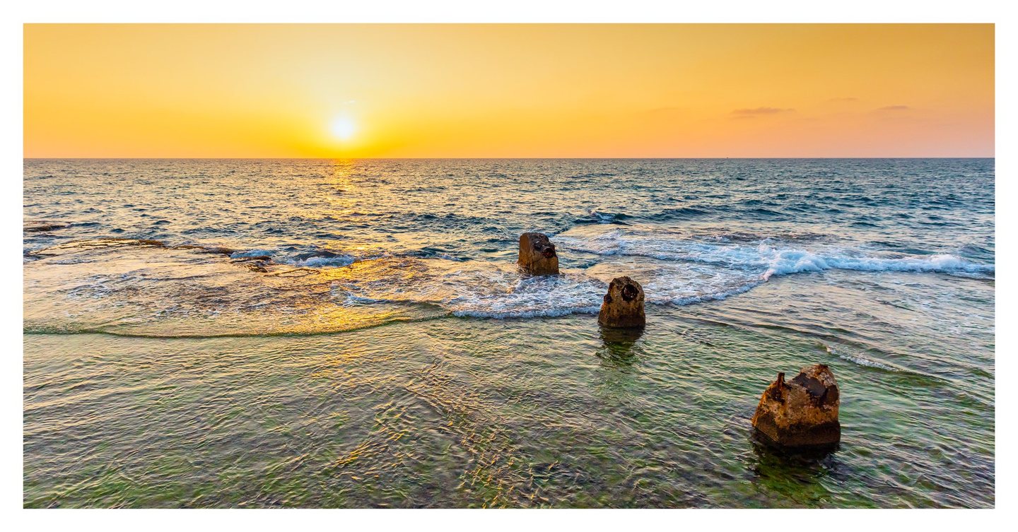 A low sun casts golden light across the sea as small waves curl around three weathered concrete remnants in clear coastal shallows.