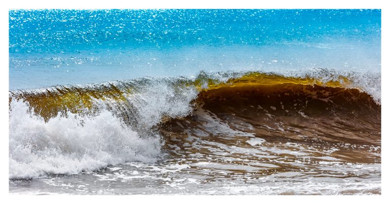 A curling amber-brown wave breaks into white foam beneath a stretch of bright turquoise water glittering in sunlight.