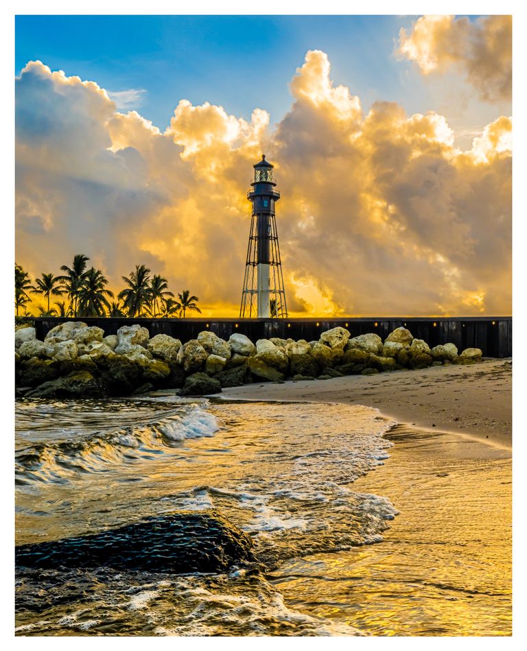 A slender lighthouse rises beside palm trees and a rocky seawall beneath golden clouds, while foamy waves wash across a sandy beach.