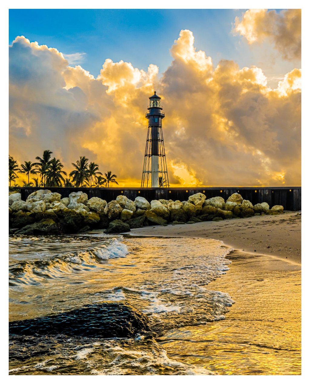 A slender lighthouse rises beside palm trees and a rocky seawall beneath golden clouds, while foamy waves wash across a sandy beach.