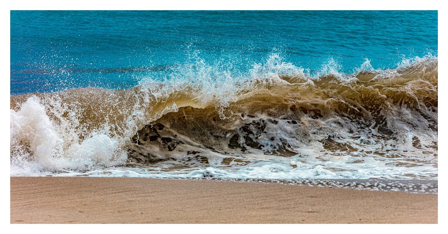 A brown-green wave breaks onto a sandy beach, throwing white foam and spray against turquoise seawater.