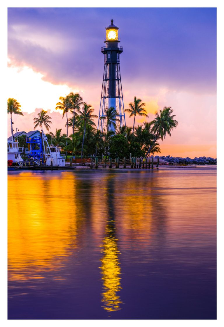 An illuminated metal lighthouse rises above palm trees and docked boats beneath a purple and pink sky, with golden reflections stretching across the water.