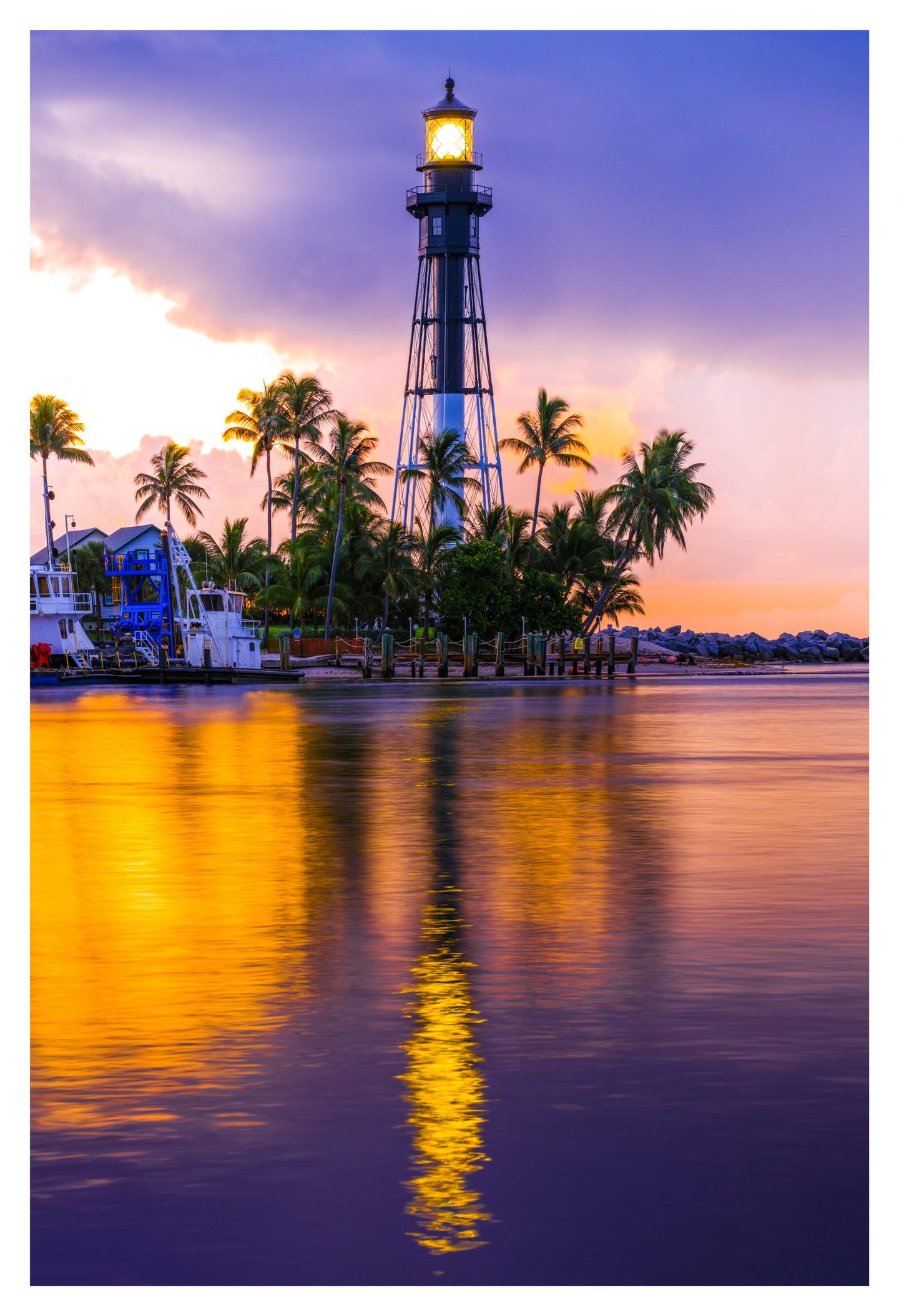 An illuminated metal lighthouse rises above palm trees and docked boats beneath a purple and pink sky, with golden reflections stretching across the water.