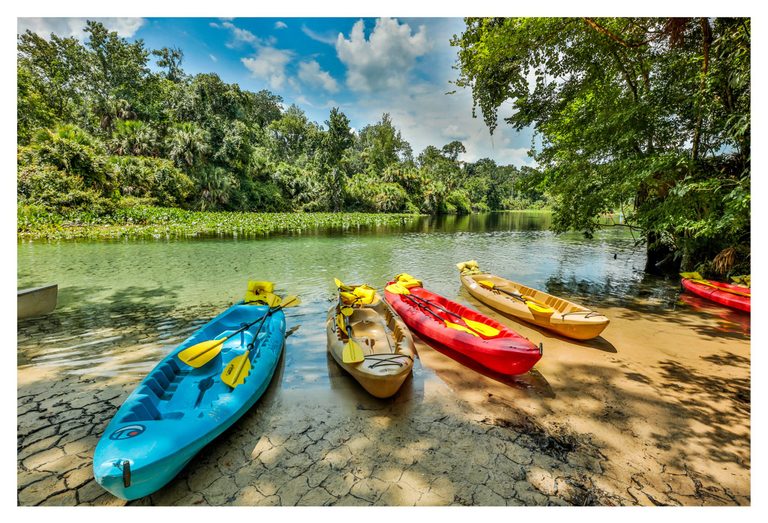 Blue, red, and tan kayaks with yellow paddles rest on a sandy riverbank beside clear green water bordered by dense trees under a partly cloudy blue sky.