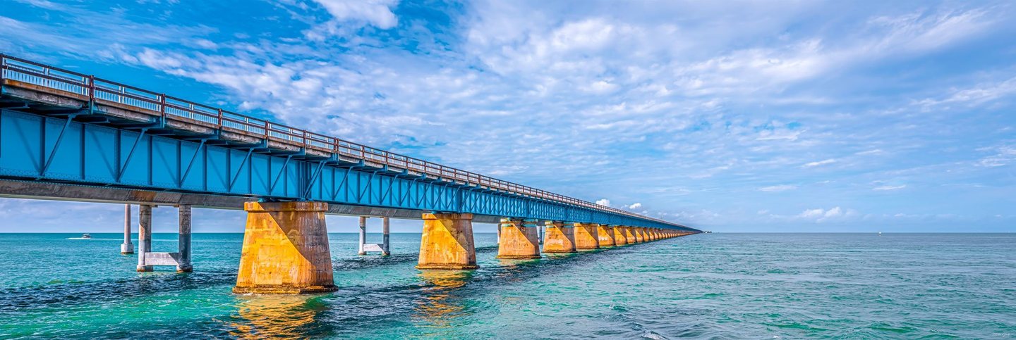 A blue steel bridge on golden concrete piers stretches across turquoise seawater toward the horizon beneath a cloud-streaked sky.