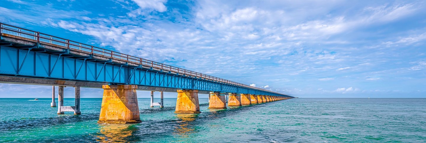 A blue steel bridge on golden concrete piers stretches across turquoise seawater toward the horizon beneath a cloud-streaked sky.