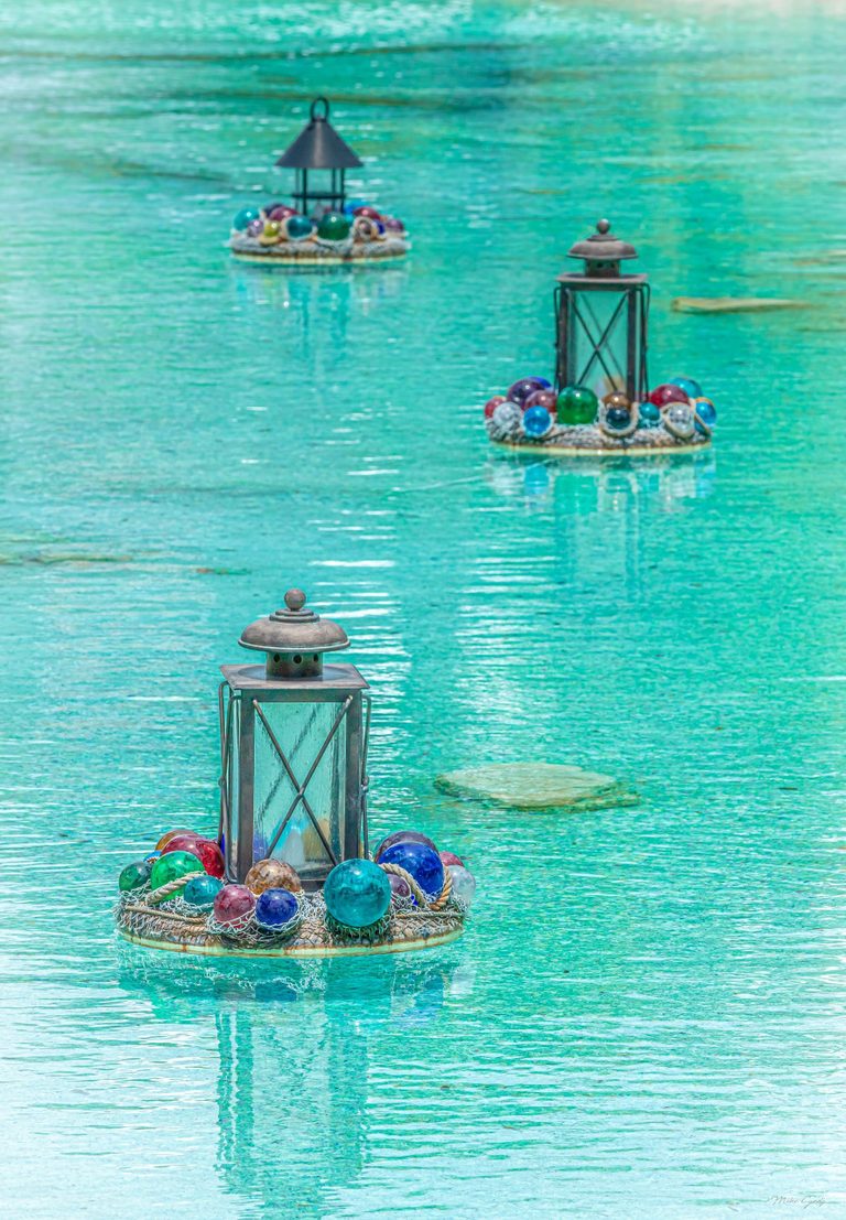 Three metal lanterns on circular floating platforms decorated with colorful glass spheres and netting recede across rippling turquoise water.