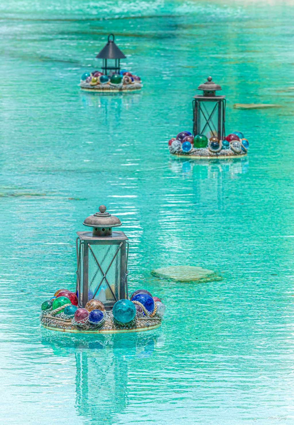 Three metal lanterns on circular floating platforms decorated with colorful glass spheres and netting recede across rippling turquoise water.