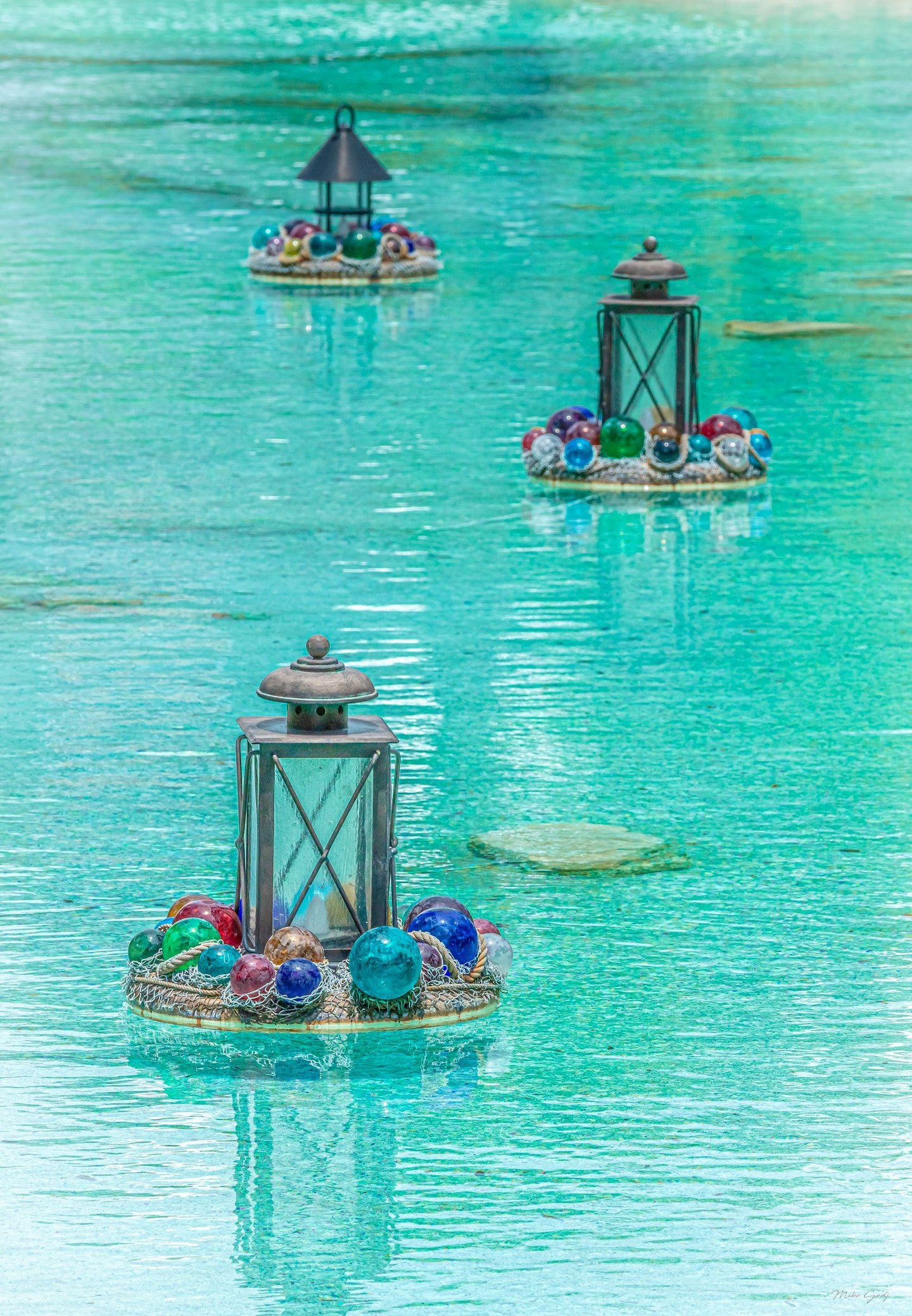 Three metal lanterns on circular floating platforms decorated with colorful glass spheres and netting recede across rippling turquoise water.