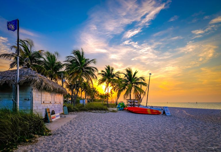 Golden light glows behind palm trees on a sandy beach, with a thatched rental hut, colorful kayaks, and calm sea beneath sweeping pink clouds.
