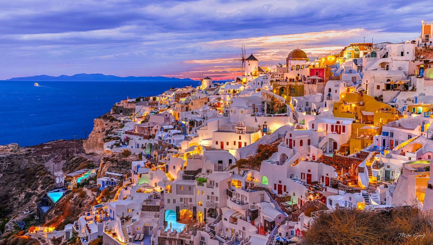 Whitewashed buildings and windmills cascade down the cliffs of Oia, Santorini, glowing with warm lights above a deep blue sea beneath a clouded sunset sky.