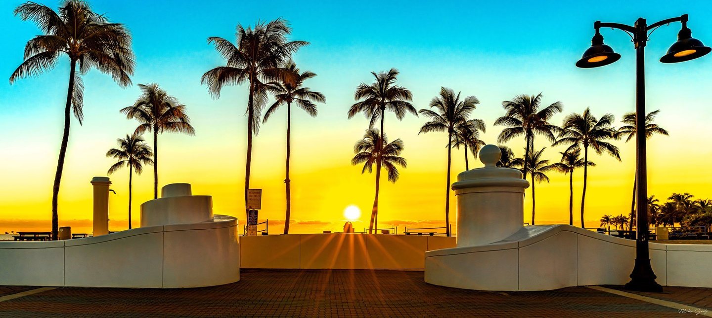 A low sun shines between palm trees along a beachfront promenade, framed by curved white walls, brick paving, and a black double streetlamp beneath a turquoise and yellow sky.