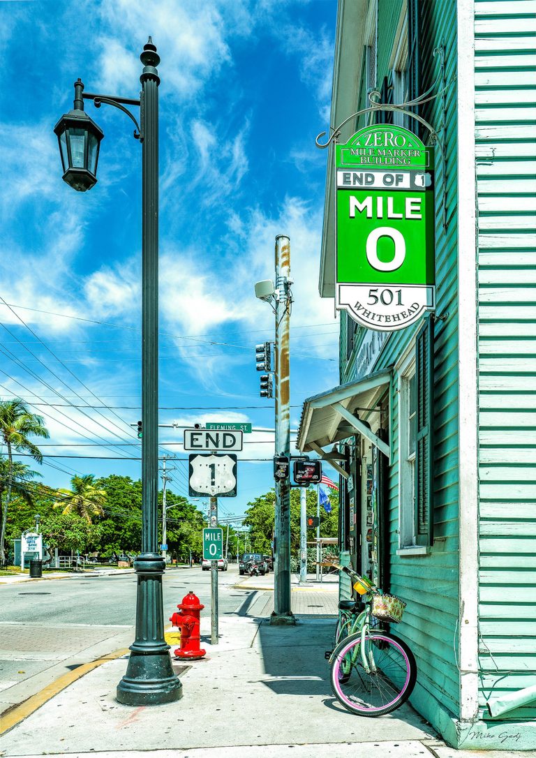 A bicycle with pink wheel rims leans against a turquoise wooden building beside Mile 0 signs, a black streetlamp, and a red fire hydrant in sunny Key West.
