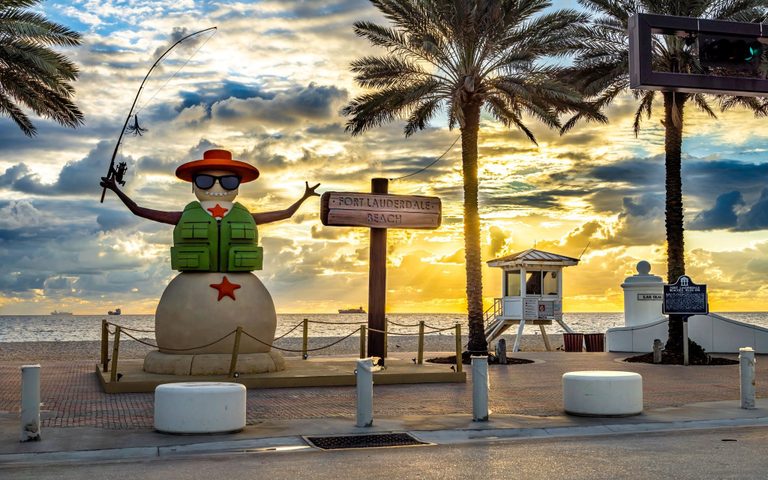A snowman sculpture wearing sunglasses and a green fishing vest holds a fishing rod beside the Fort Lauderdale Beach sign, with palms, a lifeguard tower, and golden clouds over the ocean.