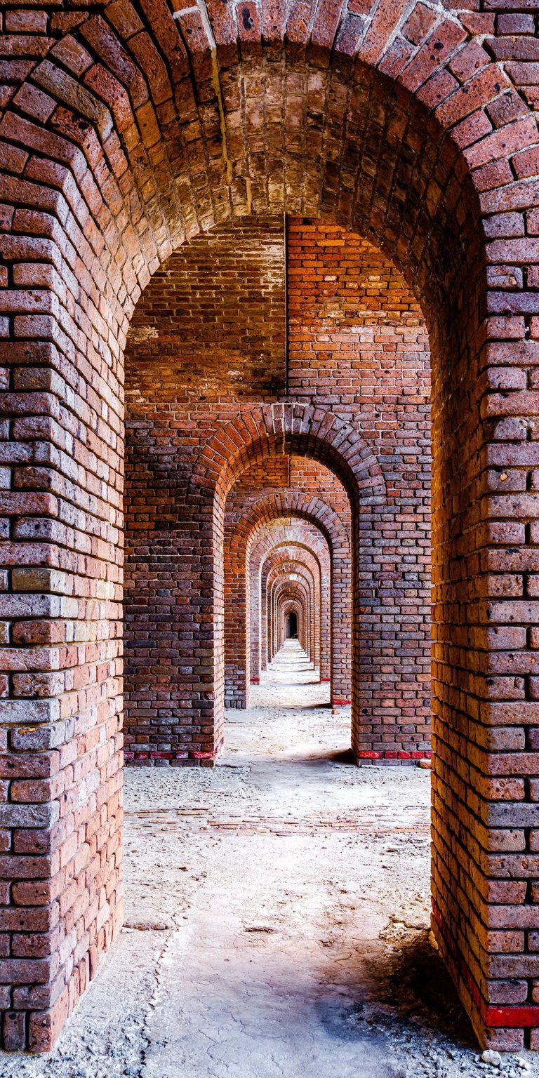 A long passage of weathered red brick arches recedes toward a dark vanishing point, with bands of light crossing the pale, uneven floor.