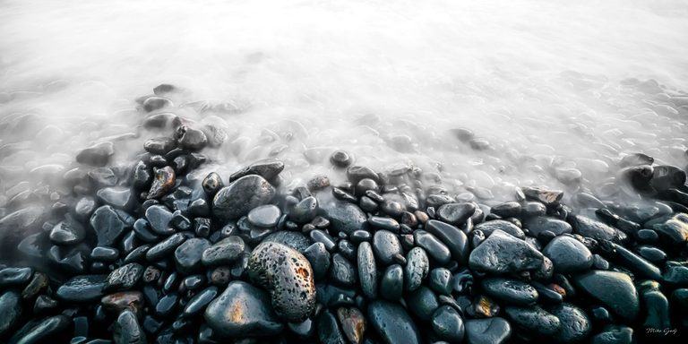 Dark wet pebbles fill the foreground of a rocky shoreline, fading beneath pale, softly blurred water toward the top of the image.