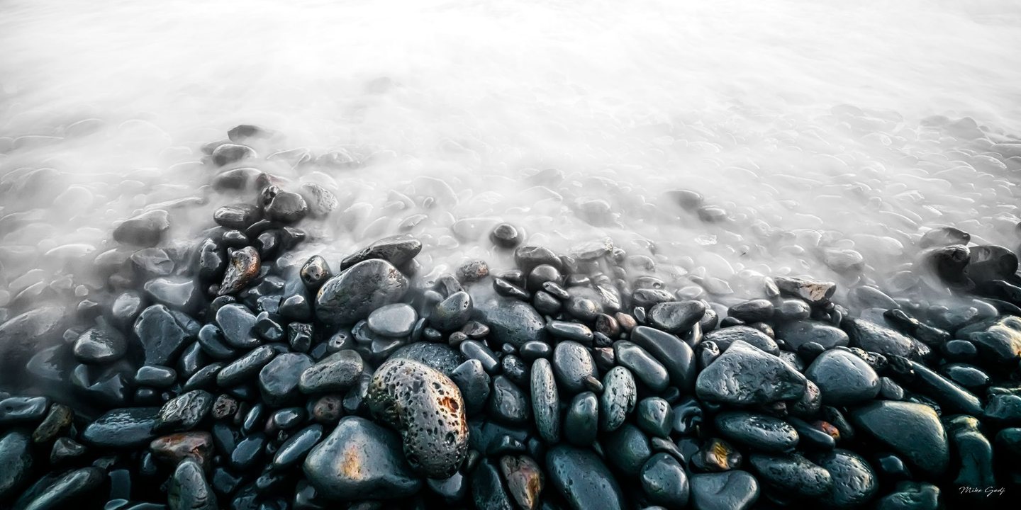 Dark wet pebbles fill the foreground of a rocky shoreline, fading beneath pale, softly blurred water toward the top of the image.