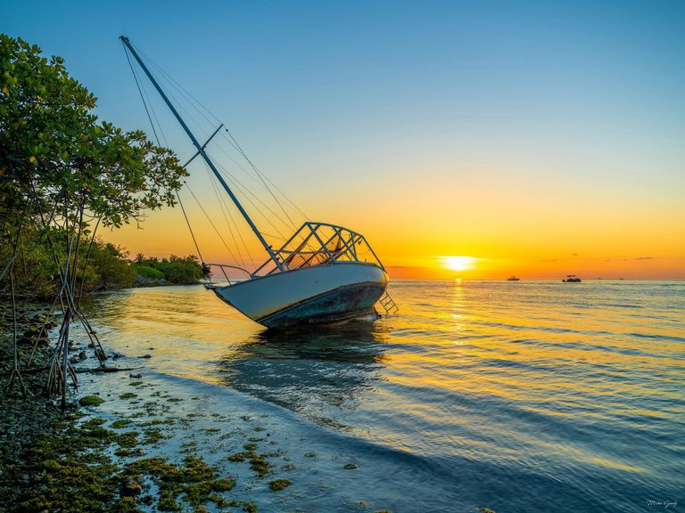 A weathered sailboat leans in shallow water beside mangroves as a golden sunset reflects across the sea, with small boats on the horizon.