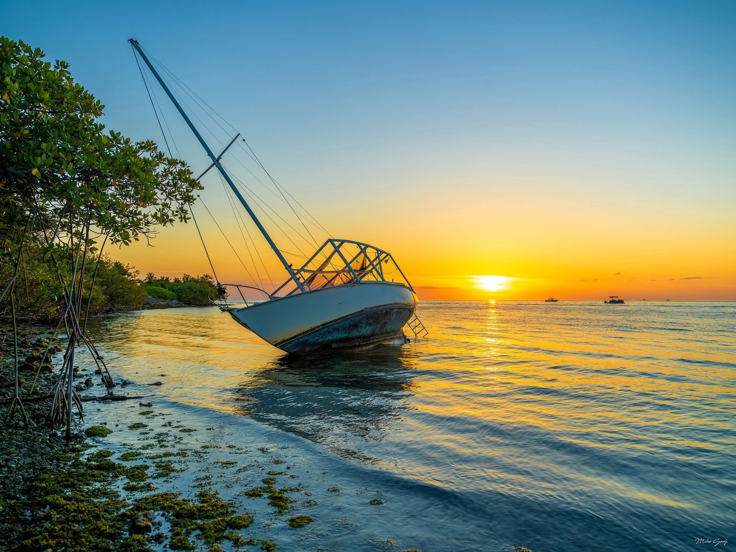 A weathered sailboat leans in shallow water beside mangroves as a golden sunset reflects across the sea, with small boats on the horizon.