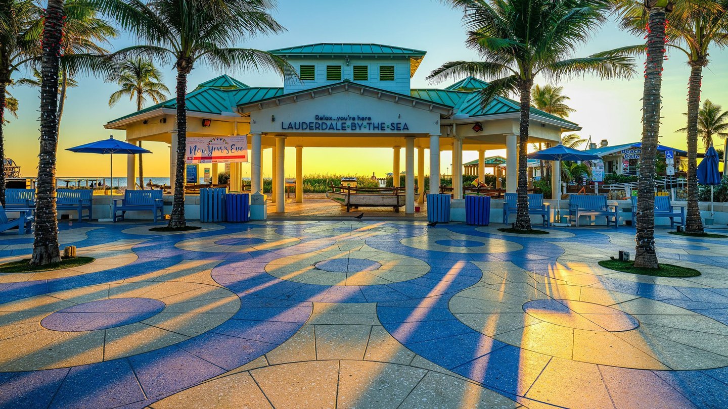 Golden light casts long palm shadows across a colorful patterned plaza in front of the turquoise-roofed beachfront pavilion at Lauderdale-by-the-Sea.