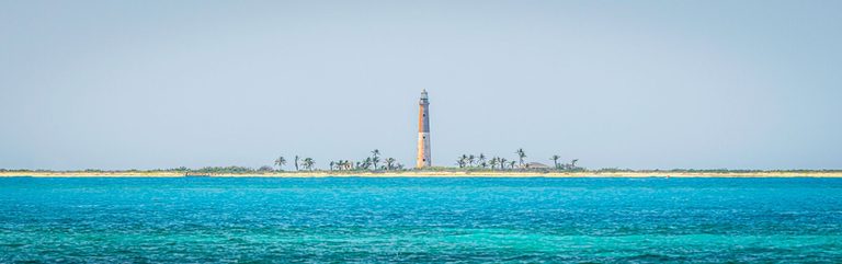A tall lighthouse rises above a low sandy island dotted with palms, across vivid turquoise water beneath a pale blue sky.