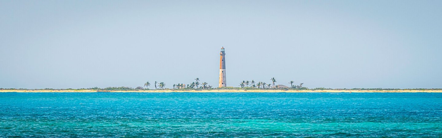 A tall lighthouse rises above a low sandy island dotted with palms, across vivid turquoise water beneath a pale blue sky.