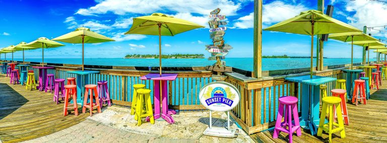 Lime-green umbrellas shade brightly painted tables and stools on Sunset Pier in Key West, overlooking turquoise water beneath a blue sky with white clouds.