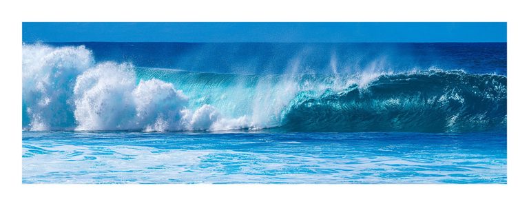 A turquoise ocean wave curls on the right and breaks into white spray on the left, with foamy water in the foreground beneath a clear blue sky.
