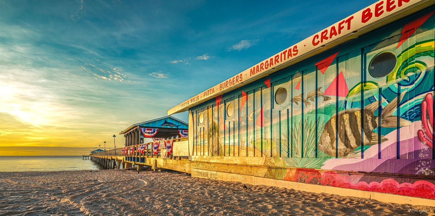 Golden light falls across a sandy beach, a pier decorated with red, white, and blue bunting, and a seaside building painted with colorful marine life.