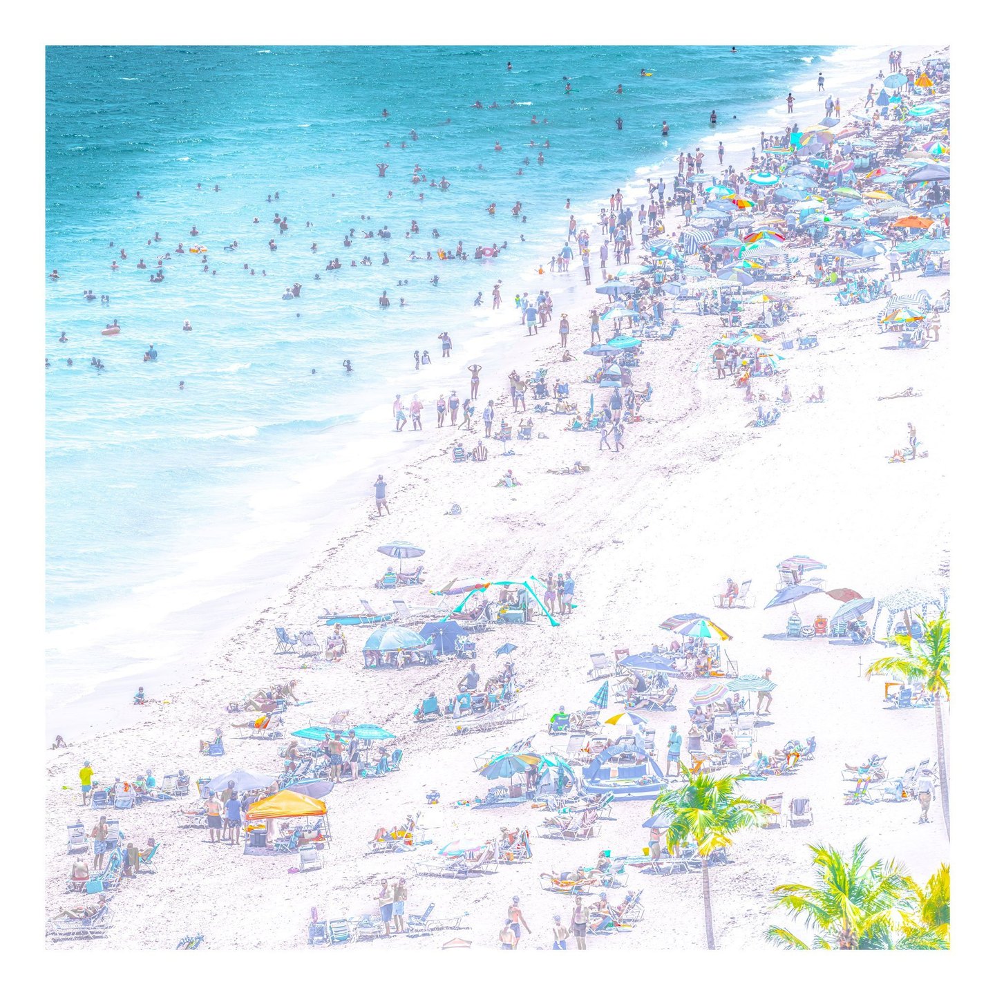 An elevated view shows swimmers in turquoise water beside a crowded pale beach scattered with colorful umbrellas, lounge chairs, and palm trees.