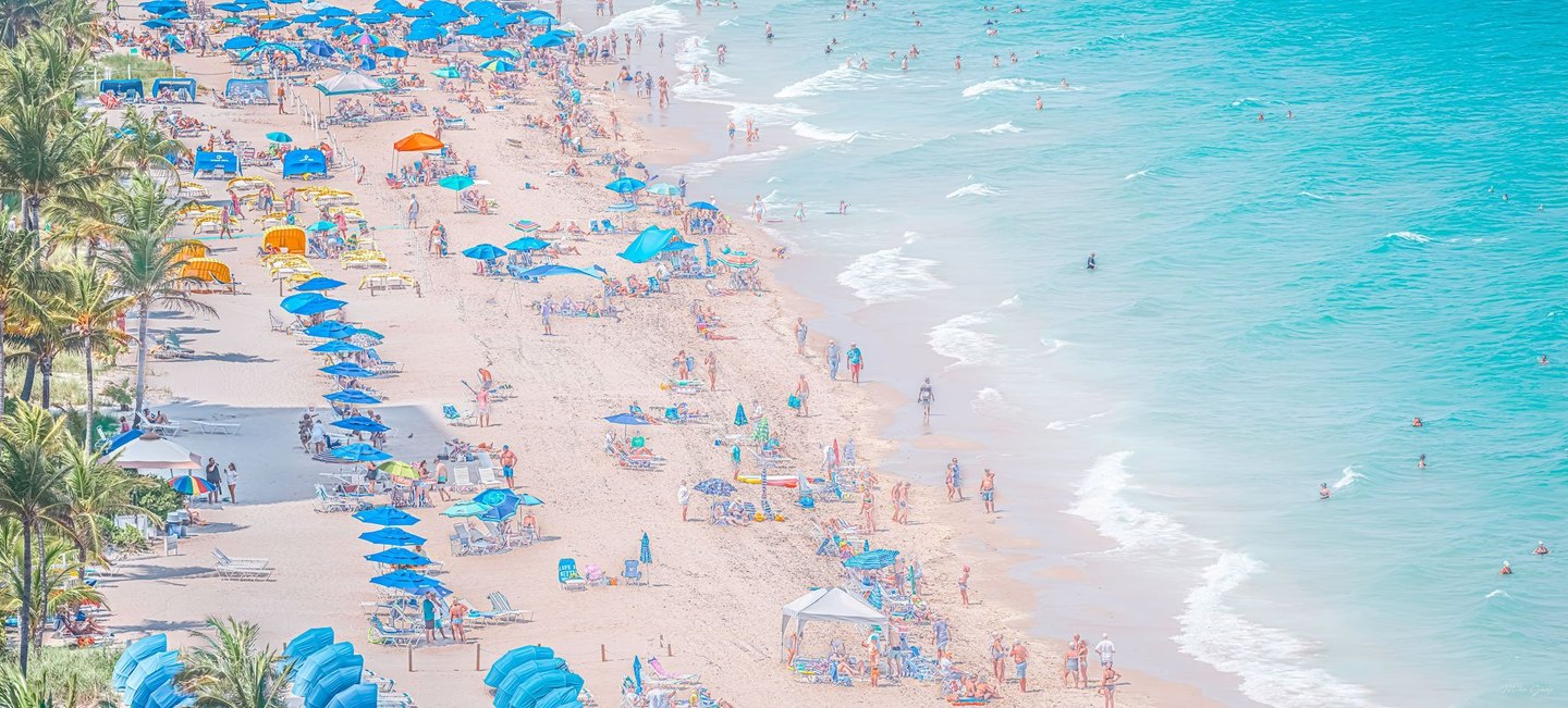 An elevated view of a crowded sandy beach shows blue umbrellas, lounge chairs and palm trees beside swimmers in turquoise water and small breaking waves.