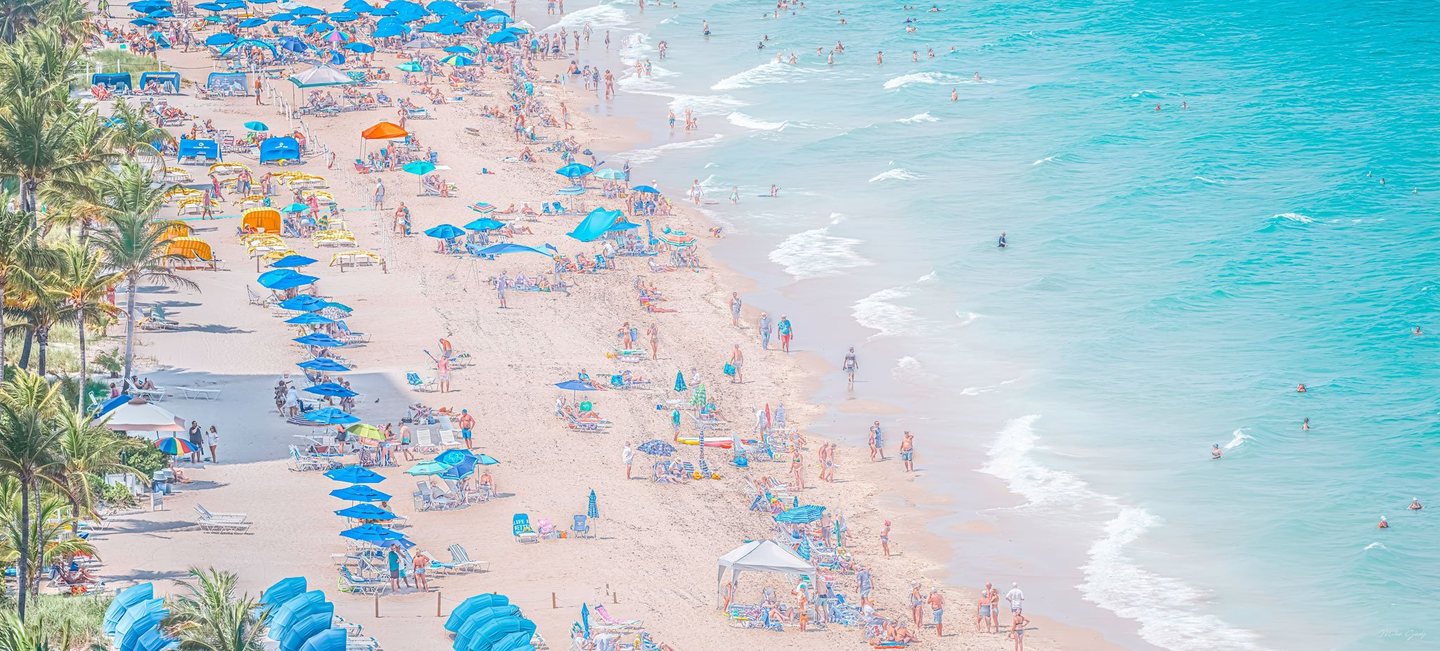 An elevated view of a crowded sandy beach shows blue umbrellas, lounge chairs and palm trees beside swimmers in turquoise water and small breaking waves.