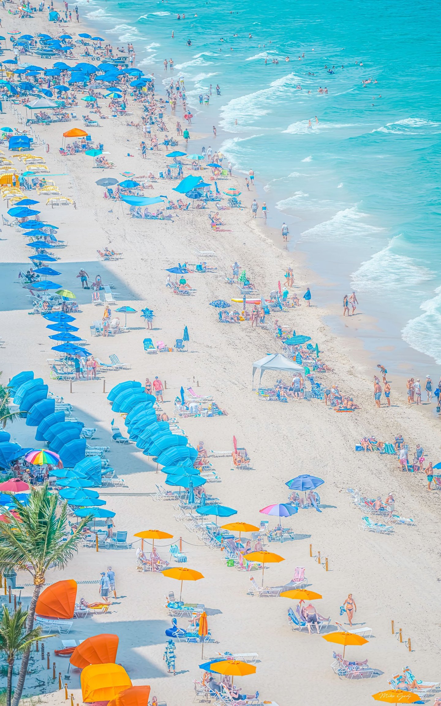 An elevated view of a busy sandy beach shows rows of blue and orange umbrellas, lounge chairs, palm trees, and swimmers in turquoise surf.