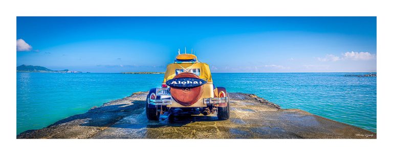 A yellow vintage-style car carrying boards and an Aloha sign is parked on a rocky point surrounded by turquoise sea beneath a clear blue sky.