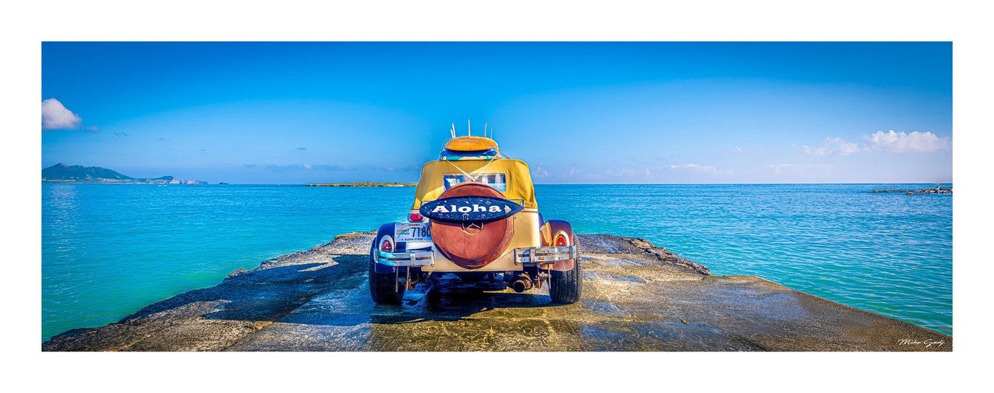 A yellow vintage-style car carrying boards and an Aloha sign is parked on a rocky point surrounded by turquoise sea beneath a clear blue sky.